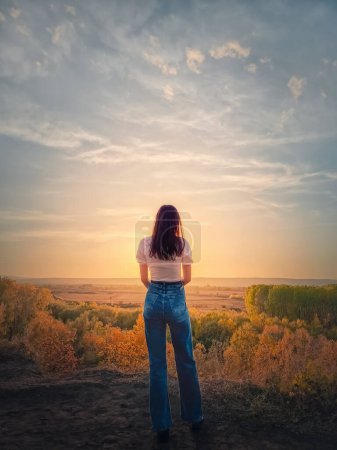 Téléchargez les photos : Vue arrière d'une jeune femme au sommet d'une colline admirant le coucher de soleil calme sur la vallée d'automne avec des arbres colorés. automne saison mélancolique sentiment, copier l'espace ciel arrière-plan - en image libre de droit