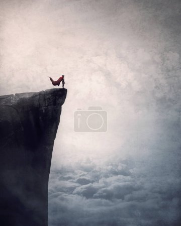 Téléchargez les photos : Superhéros avec cap se dresse courageux sur le bord d'une falaise au-dessus des nuages à la recherche déterminée à l'horizon avec la main jusqu'au front. Puissant héros scène conceptuelle - en image libre de droit
