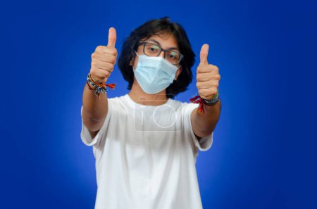Foto de Adolescente con pulgares arriba lleva una máscara médica y una camisa blanca sobre un fondo azul - Imagen libre de derechos