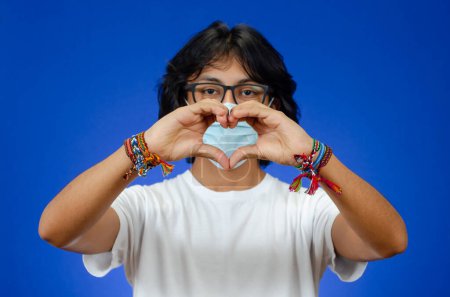 Foto de Un joven con las manos en forma de corazón lleva una máscara médica y una camisa blanca sobre fondo azul - Imagen libre de derechos