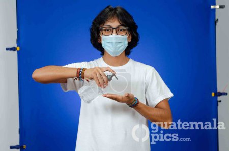 Téléchargez les photos : Jeune Latino homme avec un masque médical et chemise blanche sur un fond bleu, met un gel antibactérien et antiseptique dans ses mains - en image libre de droit