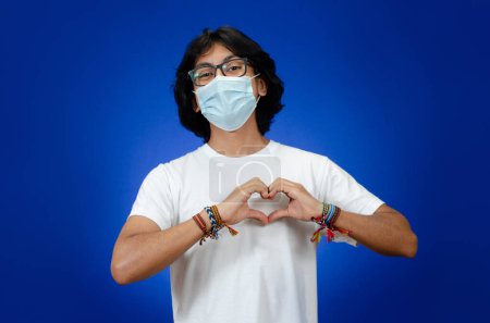Foto de Joven feliz con las manos en forma de corazón en el pecho, lleva una máscara médica y una camisa blanca - Imagen libre de derechos