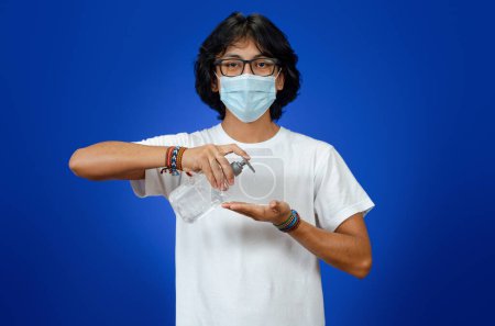 Foto de Joven latino con máscara médica y camisa blanca sobre fondo azul, se pone un gel antibacteriano y antiséptico en las manos - Imagen libre de derechos