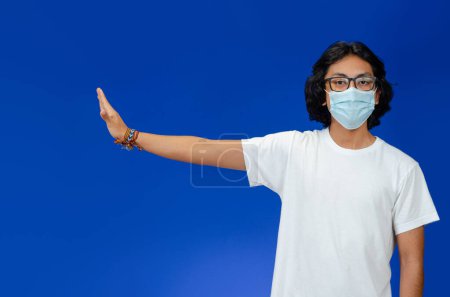 Foto de El joven hace un gesto de alto, con una máscara médica, y una camiseta blanca sobre un fondo azul - Imagen libre de derechos