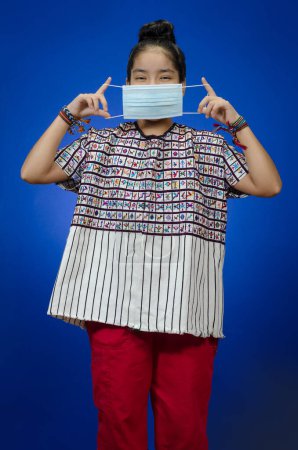 Photo for Young happy girl in a typical dress, gipil, putting on a medical mask on blue background. - Royalty Free Image