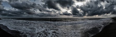 Foto de Atardecer temible clima panorámico en la playa con cielo dramático con nubes de cúmulos oscuros y mar tormentoso con olas rompiendo en la orilla generando espuma blanca. - Imagen libre de derechos