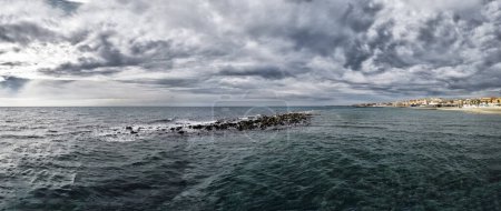Foto de Vista panorámica del mal tiempo y el cielo dramático sobre la costa romana en Ostia Lido con horizonte de la ciudad y el mar agitado mientras tanto un cuervo descansa sobre la estatua de Neptuno. - Imagen libre de derechos