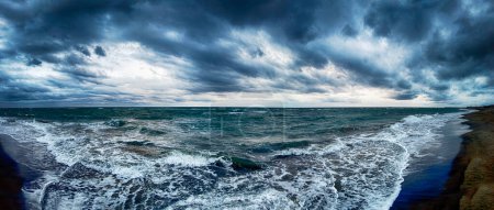 Foto de Dramático cielo nublado con nubes nimbostratus y mal tiempo en vista panorámica en la playa con olas de mar tormentosas amenazando con estrellarse en la orilla con mucha espuma de mar. - Imagen libre de derechos