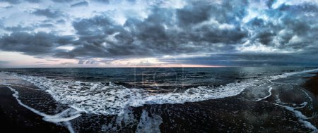 Foto de Impresionante panorámica 180 grados de mar agitado al atardecer en la hora azul en la playa, un cielo dramático con increíble luz y reflejos de nubes y sol con borde de agua estrellándose con espuma. - Imagen libre de derechos