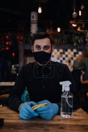 Foto de Young restaurant waiters cleaning and disinfecting tables and surfaces during Coronavirus pandemic disease. They are wearing protective face masks and gloves. - Imagen libre de derechos