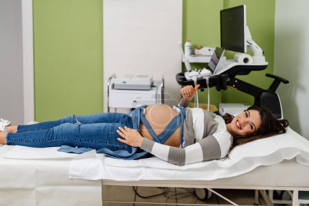 Téléchargez les photos : Belle jeune femme enceinte à l'électrocardiographe vérifier la santé du bébé et la date de naissance prévue. Surveillance cardiaque du fœtus. Concept de soins de santé et de services médicaux. - en image libre de droit
