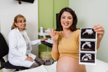 Téléchargez les photos : Beautiful and pregnant woman is happy after pregnancy medical check up. She is holding and looking at ultrasound scan of her baby and looking at camera. Modern pregnancy healthcare concept. - en image libre de droit