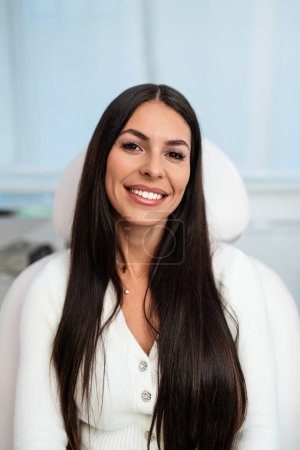 Foto de Beautiful and happy young woman sitting in medical chair and looking at camera. She is satisfied after successful beauty treatment with hyaluronic acid fillers or botulinum toxin injections. - Imagen libre de derechos