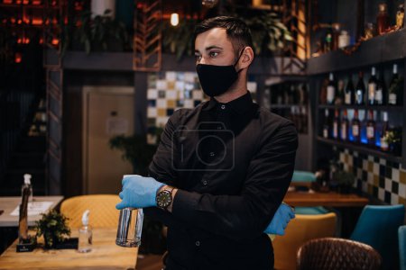 Foto de Retrato de un camarero serio parado en un bonito restaurante. Lleva una máscara protectora y guantes como parte de las medidas de seguridad contra la pandemia del Coronavirus.. - Imagen libre de derechos