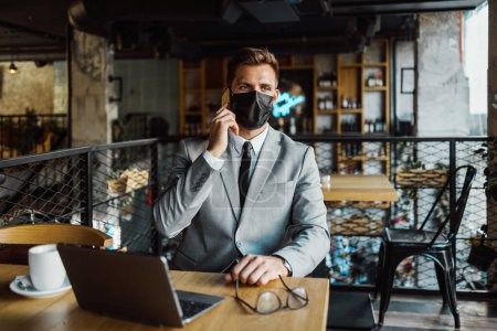 Foto de Un hombre de negocios con una máscara protectora en la cara está sentado en un café y hablando por teléfono mientras usa la computadora portátil. Coronavirus, concepto Covid-19. - Imagen libre de derechos