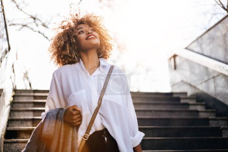 Beautiful and successful black woman walking on street. She is happy and looks up with head raised on her way to work or home. Bright sunny day and real warm flare.