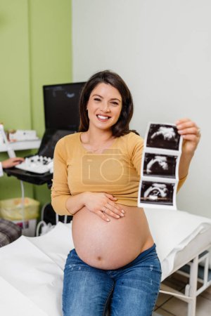 Téléchargez les photos : Belle et enceinte femme est heureuse après la grossesse examen médical. Elle tient et regarde l'échographie de son bébé et regarde la caméra. Concept moderne de soins de grossesse. - en image libre de droit