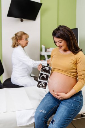 Téléchargez les photos : Belle et enceinte femme est heureuse après la grossesse examen médical. Elle tient et regarde l'échographie de son bébé. Concept moderne de soins de grossesse. - en image libre de droit