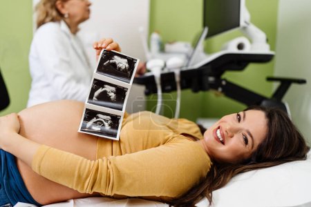 Téléchargez les photos : Belle et enceinte femme est heureuse après la grossesse examen médical. Elle tient et regarde l'échographie de son bébé et regarde la caméra. Concept moderne de soins de grossesse. - en image libre de droit