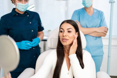 Photo for Beautiful and happy young woman sitting in medical chair and looking in the mirror. She is satisfied after successful beauty treatment with hyaluronic acid fillers or botulinum toxin injections. - Royalty Free Image