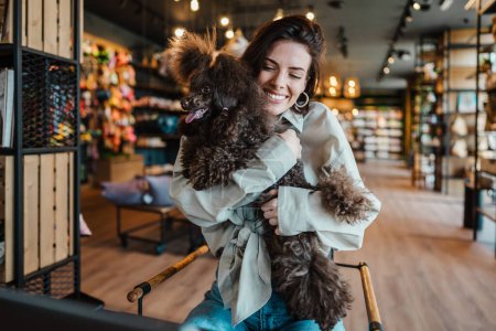 Foto de Hermosa y feliz joven sentada en el moderno bar de la tienda de mascotas y disfrutando en café fresco junto con su adorable caniche de juguete marrón. - Imagen libre de derechos