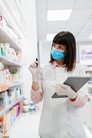 Téléchargez les photos : Magnifique pharmacien avec masque protecteur qui travaille et se tient debout dans une pharmacie et fait un bilan. Portrait d'un travailleur de la santé positif ou d'un chimiste à son travail. - en image libre de droit