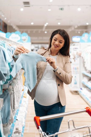 Téléchargez les photos : Beautiful young pregnant mother choosing and buying colorful clothes and appliances for her new incoming baby in. Child shop or store concept. - en image libre de droit