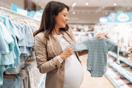 Photo for Beautiful young pregnant mother choosing and buying colorful clothes and appliances for her new incoming baby in. Child shop or store concept. - Royalty Free Image