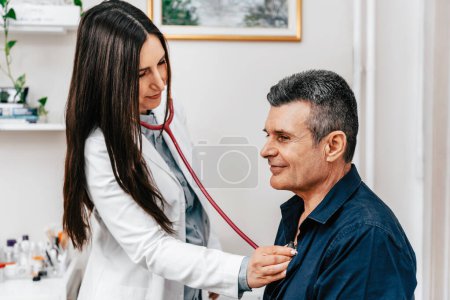 Photo for Experienced female doctor examining an male elderly patient with a stethoscope in a modern and bright doctor's office. - Royalty Free Image