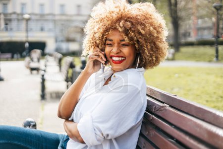 Foto de Hermosa mujer negra se sienta en el banco de la calle y utiliza el teléfono inteligente para la comunicación en línea. Ella es feliz y sonrió. Día soleado brillante. - Imagen libre de derechos