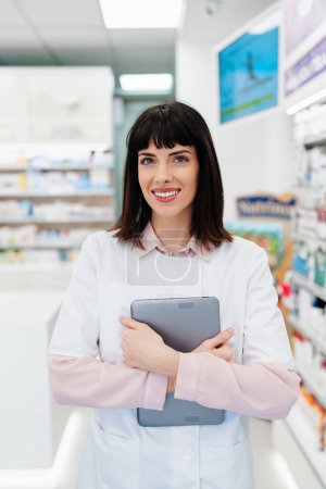 Téléchargez les photos : Magnifique pharmacien travaillant et se tenant dans une pharmacie et faisant un bilan. Portrait d'un travailleur de la santé positif ou d'un chimiste à son travail. Elle est debout et regarde la caméra. - en image libre de droit