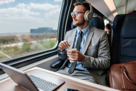 Foto de Hombre de negocios guapo se está divirtiendo mientras viaja en tren de alta velocidad. Él está utilizando ordenador portátil y auriculares inalámbricos para la comunicación en línea, juegos y entretenimiento. - Imagen libre de derechos