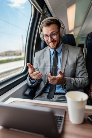 Foto de Hombre de negocios guapo se está divirtiendo mientras viaja en tren de alta velocidad. Él está utilizando ordenador portátil y auriculares inalámbricos para la comunicación en línea, juegos y entretenimiento. - Imagen libre de derechos