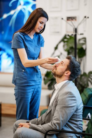 Schöne Medizintechnikerin, die vor einer Augenuntersuchung einem stattlichen männlichen Patienten Pupillen-Dilatationstropfen aufträgt. Moderne Klinik zur Diagnose und Behandlung von Augen- und Seherkrankungen.