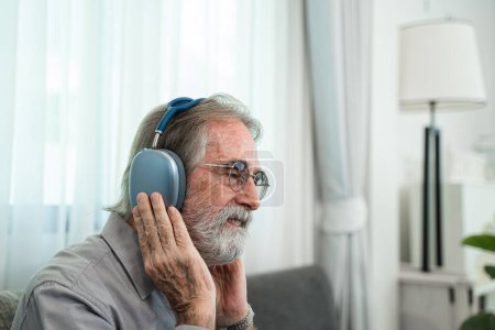 Téléchargez les photos : Homme âgé écoutant de la musique avec écouteurs, Détendu dans le canapé à sa maison. - en image libre de droit