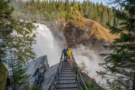 Téléchargez les photos : Couple debout sur un pont de belvédère à la célèbre cascade de Tannforsen, au nord de la Suède, avec un arc-en-ciel dans la brume et des cascades d'eau rapides. - en image libre de droit