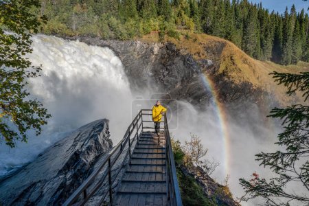 Photo for Girls standing on viewpoint bridge at Famous waterfall Tannforsen northern Sweden, with a rainbow in the mist and rapid flowing cascades of water. - Royalty Free Image