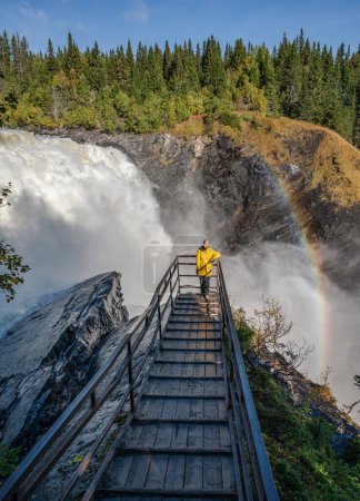 Foto de Chicas de pie en el puente de mirador en la famosa cascada Tannforsen norte de Suecia, con un arco iris en la niebla y las cascadas de agua de flujo rápido. - Imagen libre de derechos