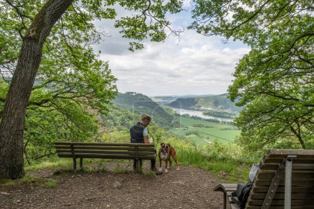 Touristenmädchen sitzt auf einer Bank mit einem Welpen-Boxerhund und betrachtet das Rheintal bei Andernach von einem Aussichtspunkt aus.