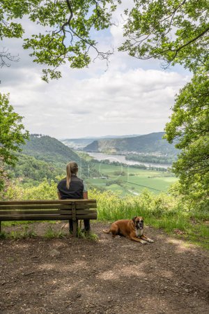 Chica turística sentada en un banco con un perro boxeador cachorro mirando el valle del río Rin cerca de Andernach desde el mirador.