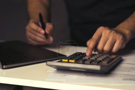 Hombre asiático está utilizando una calculadora para calcular sus gastos varios mensuales en casa.