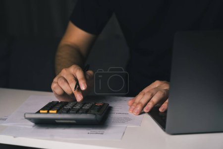 Téléchargez les photos : Asiatique homme utilise une calculatrice pour calculer les dépenses mensuelles diverses de sa famille à la maison et les enregistrer dans son ordinateur portable. - en image libre de droit