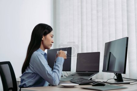 Téléchargez les photos : Femme asiatique buvait du café tôt le matin tout en contemplant le programme et le code sur l'écran de l'ordinateur. - en image libre de droit