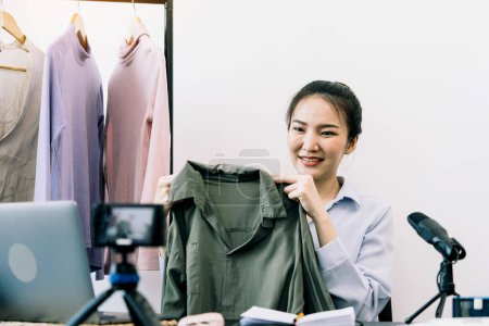Téléchargez les photos : Les jeunes entrepreneurs asiatiques ramassent leurs vêtements pour montrer aux clients grâce à des ventes en ligne sur un téléphone en direct. - en image libre de droit