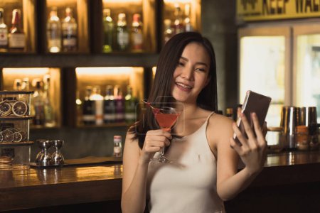 Téléchargez les photos : Femme asiatique prendre une photo d'elle-même tout en buvant du whisky au bar. - en image libre de droit