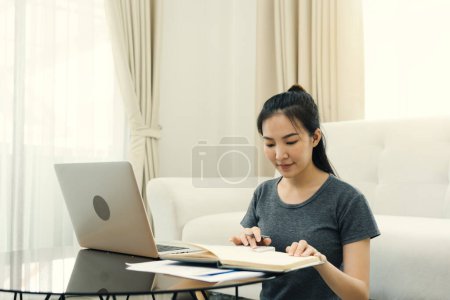 Téléchargez les photos : Jeune femme asiatique est assis dans le salon à l'aide d'une calculatrice pour calculer les revenus et les dépenses de la famille et l'écrit dans son ordinateur portable ou portable à la maison. - en image libre de droit