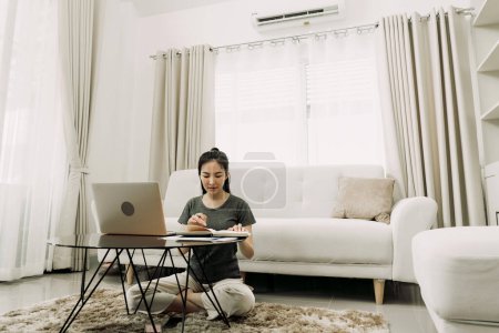 Téléchargez les photos : Jeune femme asiatique est assis dans le salon à l'aide d'une calculatrice pour calculer les revenus et les dépenses de la famille et l'écrit dans son ordinateur portable ou portable à la maison. - en image libre de droit