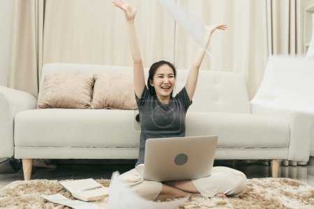 Téléchargez les photos : Asiatique femme jette des papiers dans le salon comme elle termine son travail avec son ordinateur portable sur ses genoux. - en image libre de droit