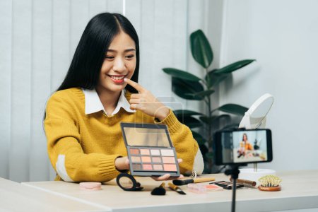 Téléchargez les photos : Jeune femme asiatique vlogging sur les produits cosmétiques soins de la peau produits sur la table avec sa caméra vidéo et démontre l'utilisation des produits et des critiques pour son canal blog en ligne. - en image libre de droit