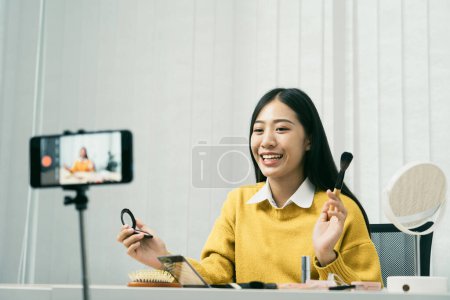 Téléchargez les photos : Jeune femme asiatique vlogging elle-même sur les produits de beauté avec faire une vidéo pour son blog sur les cosmétiques en utilisant la caméra de téléphone à la maison. - en image libre de droit